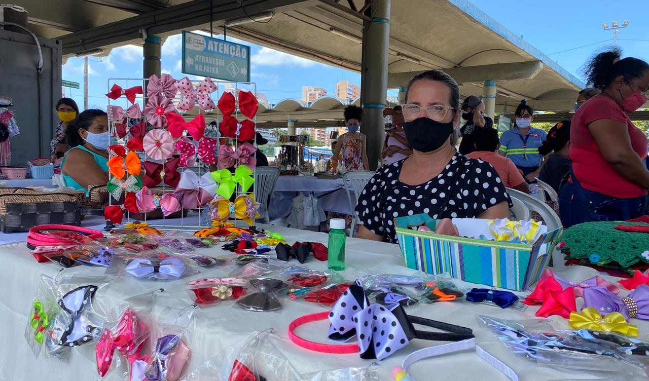 mulher sentada à frente de uma mesa dentro de um terminal de ônibus; na mesa estão colocadas tiaras de cabelo com laços coloridos